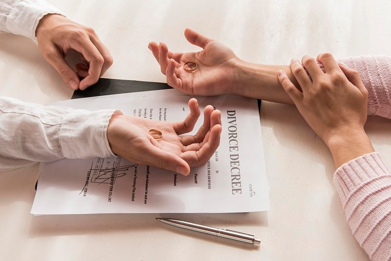 Two hands holding wedding rings over a divorce decree document with a pen beside it, symbolizing the end of a marriage.