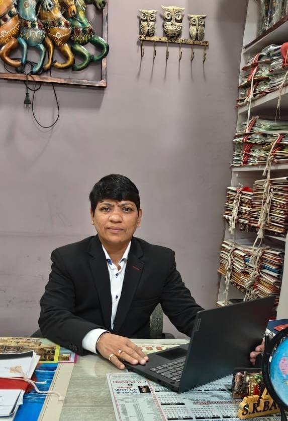 SR Baghel sitting at a desk with a laptop, surrounded by files and legal documents, in an office with decorative wall hangings and shelves.